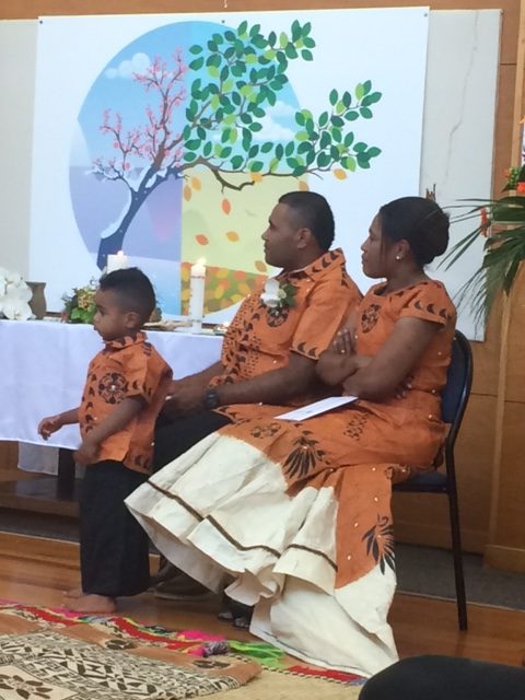 Young Bati stands in front of his seated parents, with a communion table and banner behind them. All are look left, and in matching orange wedding clothes, with Fijian patterns in black.